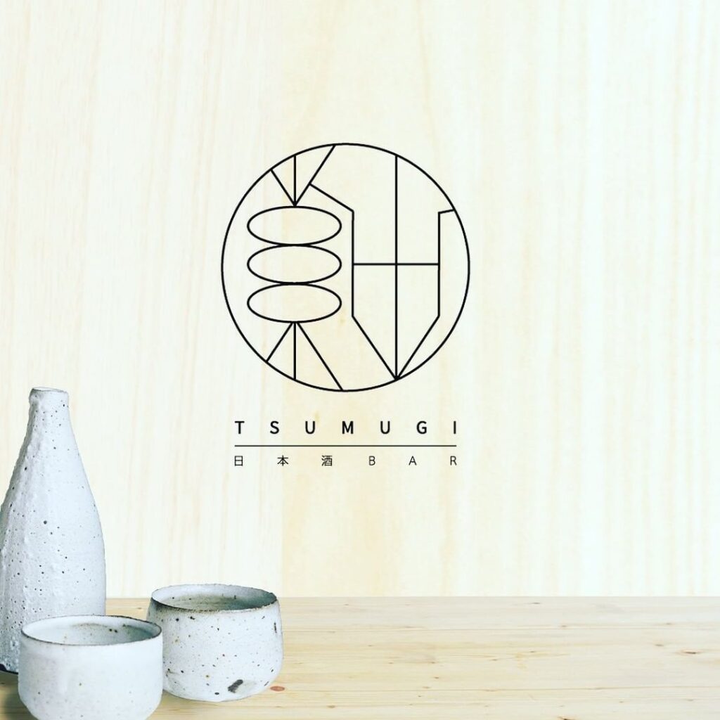 日本酒BAR TSUMUGIのロゴと、木目背景の和モダンな雰囲気に置かれたとっくりとおちょこ