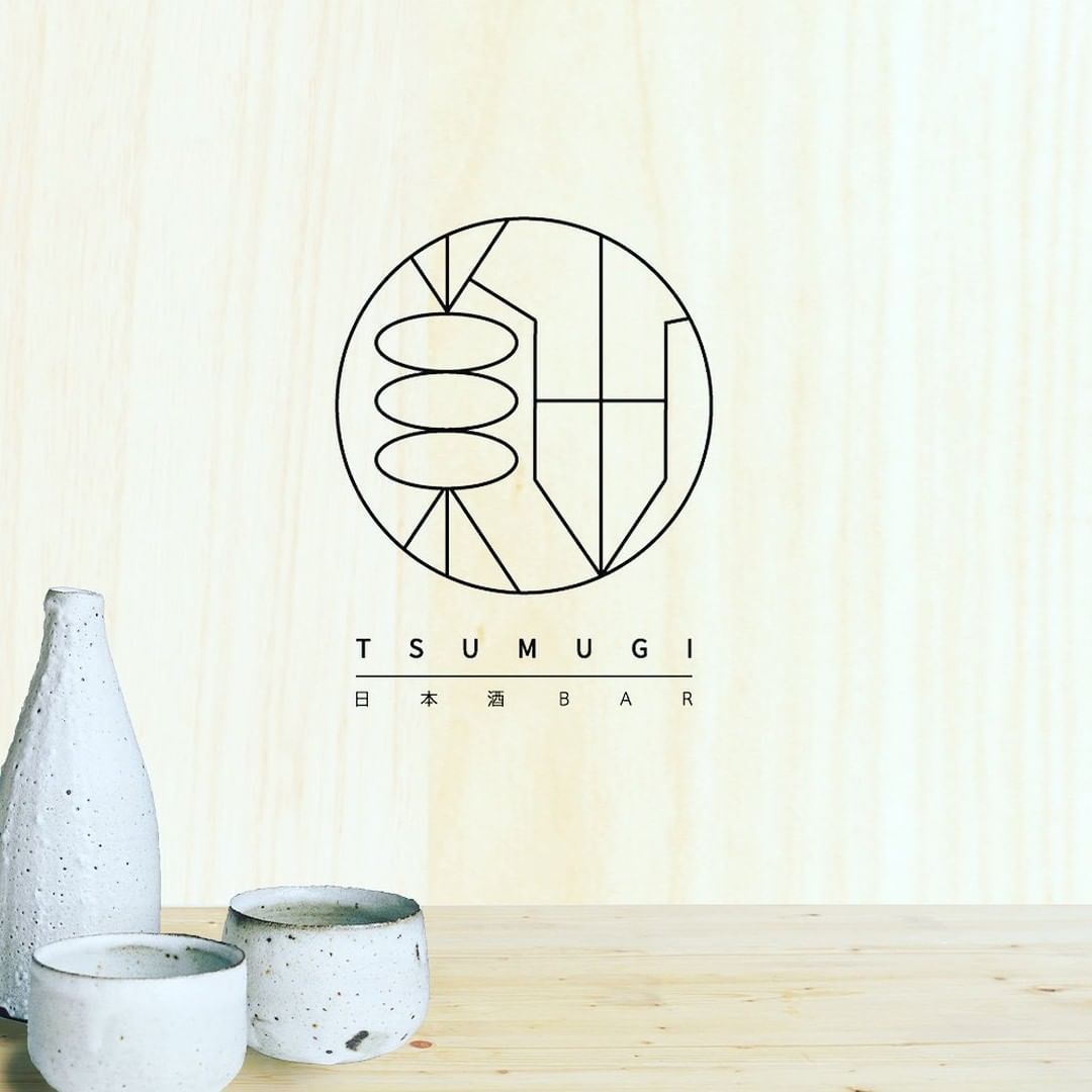 日本酒BAR TSUMUGIのロゴと、木目背景の和モダンな雰囲気に置かれたとっくりとおちょこ